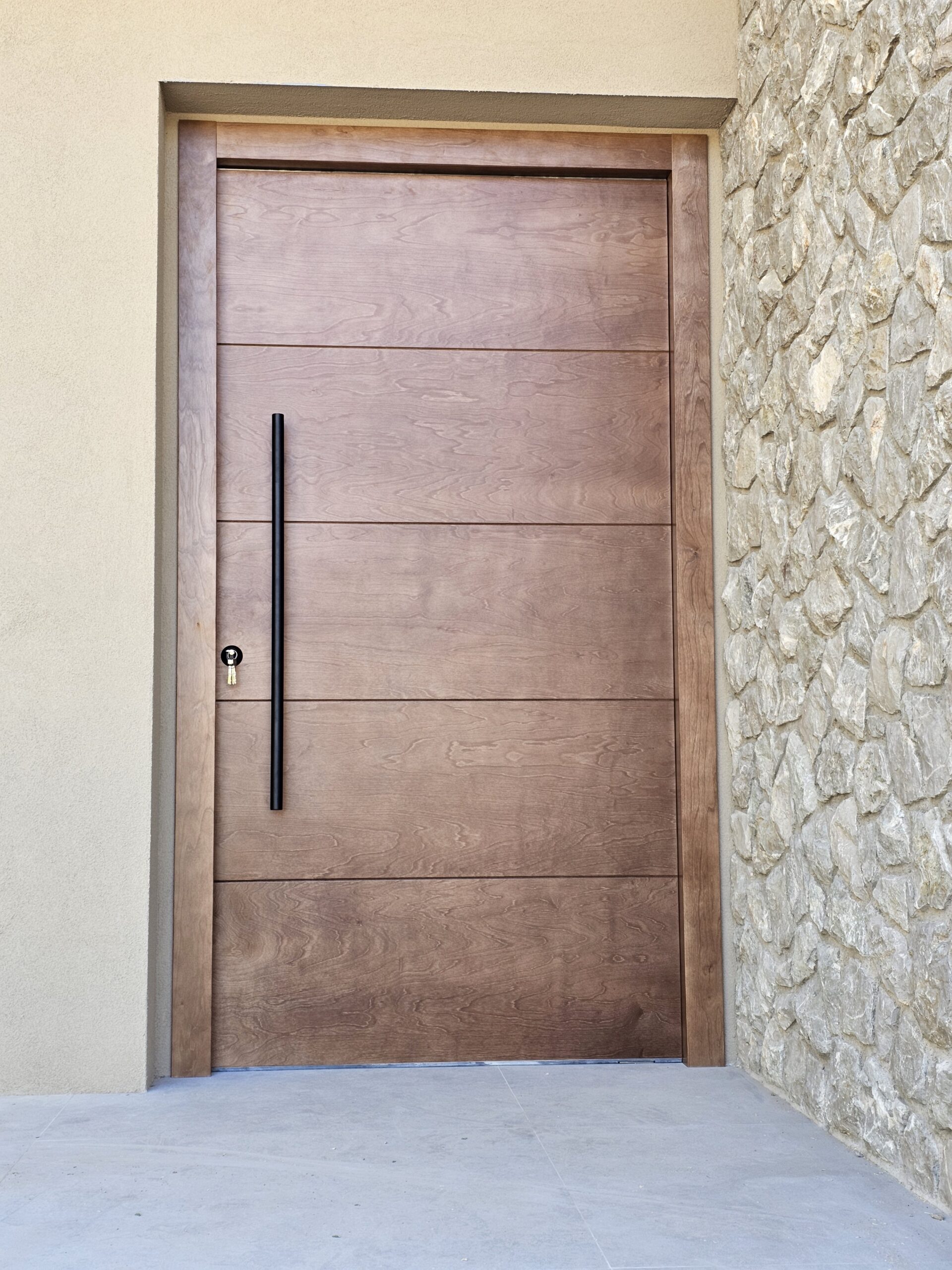 puerta-entrada-martorell Puerta de entrada de madera con tirador negro vertical junto a pared de piedra.